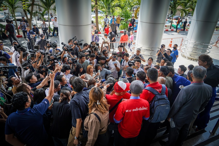 A huge pack of media point cameras and microphones at a man outside a building.