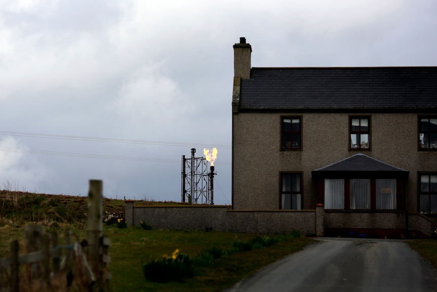 A barren, brown brick home in the foreground with a gas flare in the background