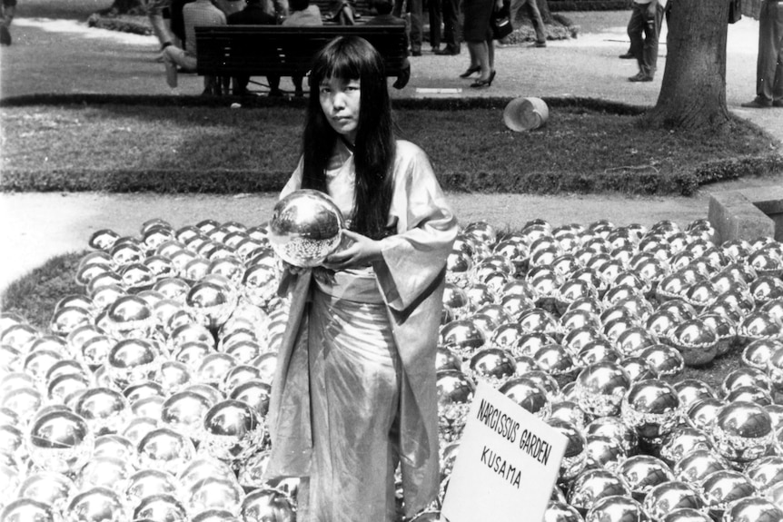 Black and white image of younger Yayoi Kusama, in flowing dress, surrounded by many small shiny balls.