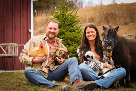 a woman and man smile as they sit beside a barn holding a chicken, pig and lamb, with a pony standing next to them 