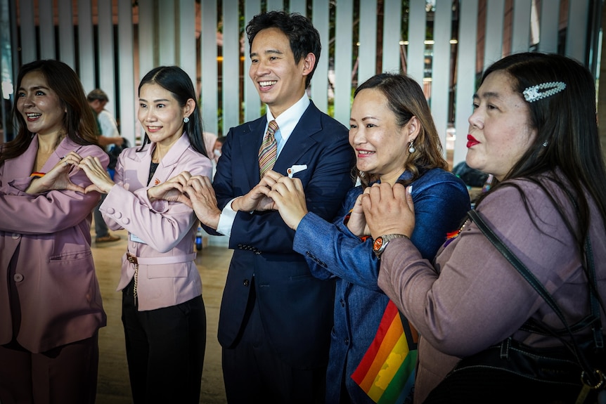 A smiling, suited male politician links hands to make heart signs with four women who stand beside him in groups of two.