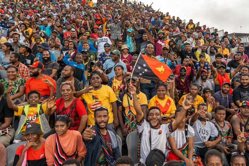 A crowd of NRL fans is pictured, they are cheering. Some hold flags and wear team jerseys.