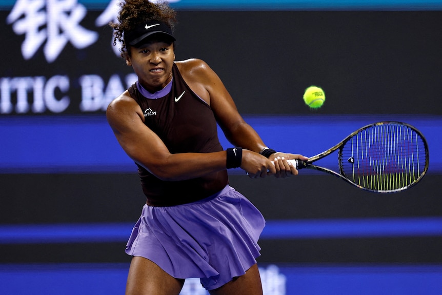 Naomi Osaka wears black and purple Nike outfit, hitting a tennis ball.