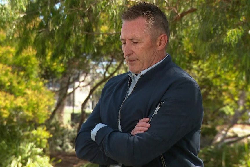 Dale Bowles wears a navy jacket over a light blue collared shirt and stands under a tree with his arms folded looking down.