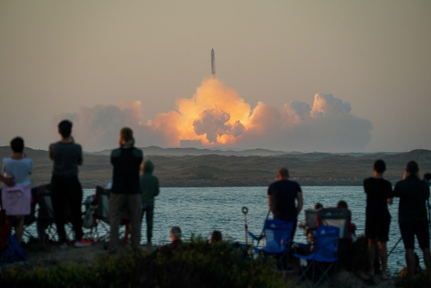 A crowd of people watch as a rocket launches out of a cloud of smoke 