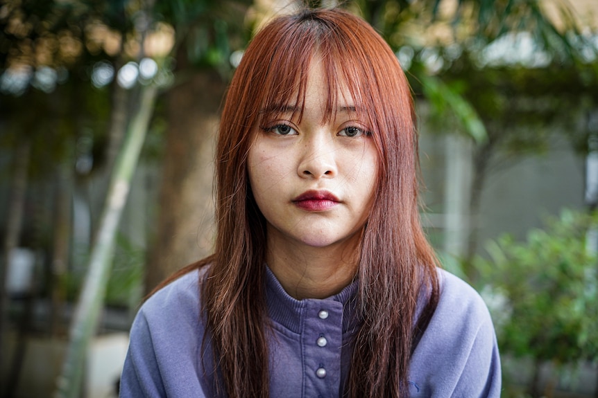 A young woman looks neutrally into the camera as she poses for a head shot outside.