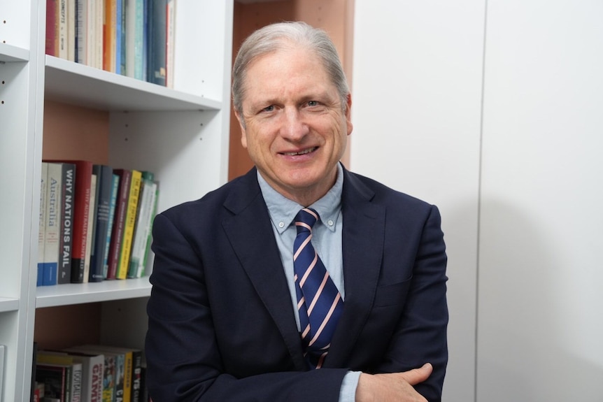 man sits in office in front of book case in suit and tie 