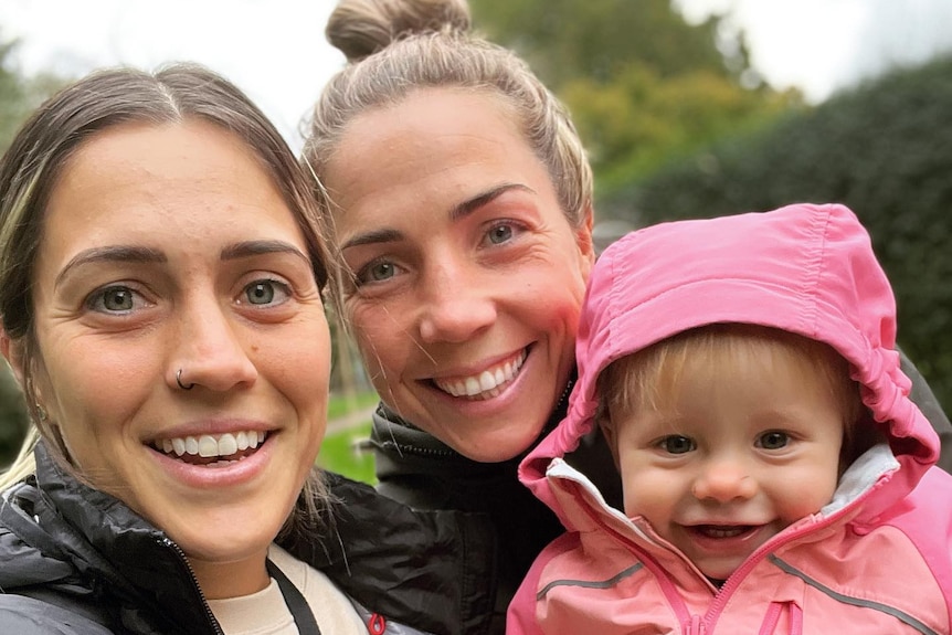 Selfie two blonde women holding a toddler girl and she wears a pink hooded jumper