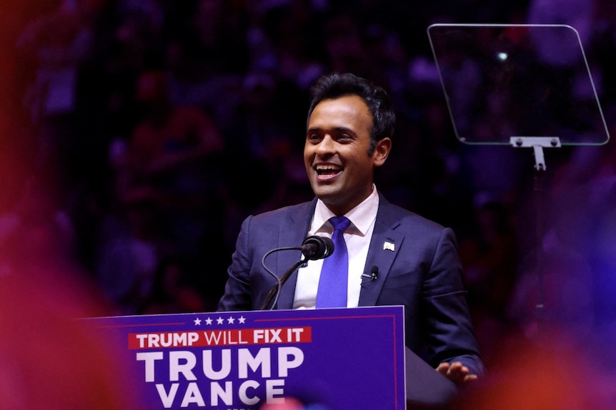 A man in a suit standing at a lecturn and speaking with a smile on his face.