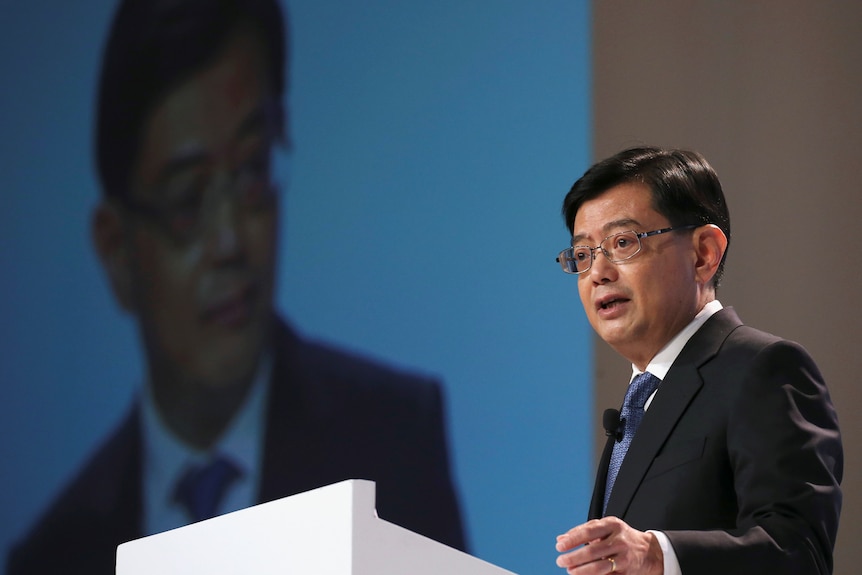 A man in a suit and glasses stands at a lectern 