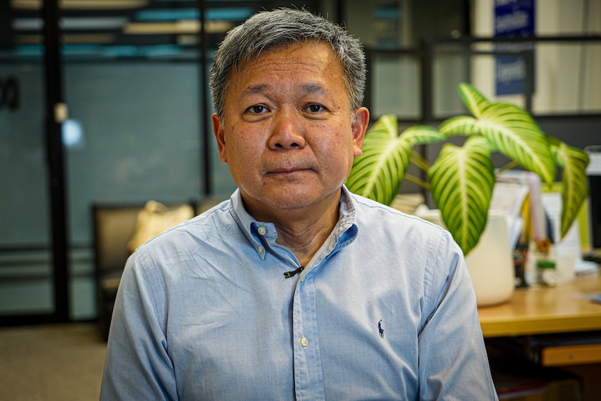 A man in a button up shirt looks into the camera as he poses for a photo inside an office.