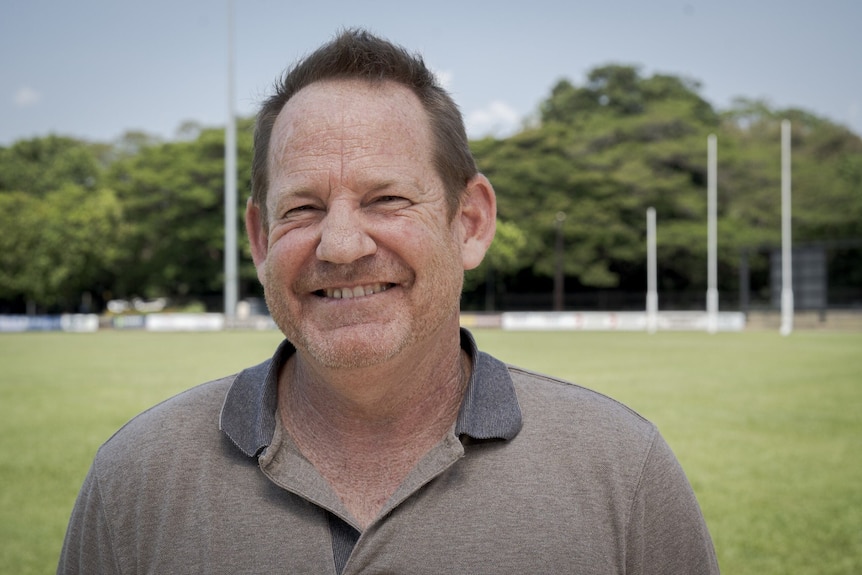 Man wearing polo shit stands at football oval and smiles 