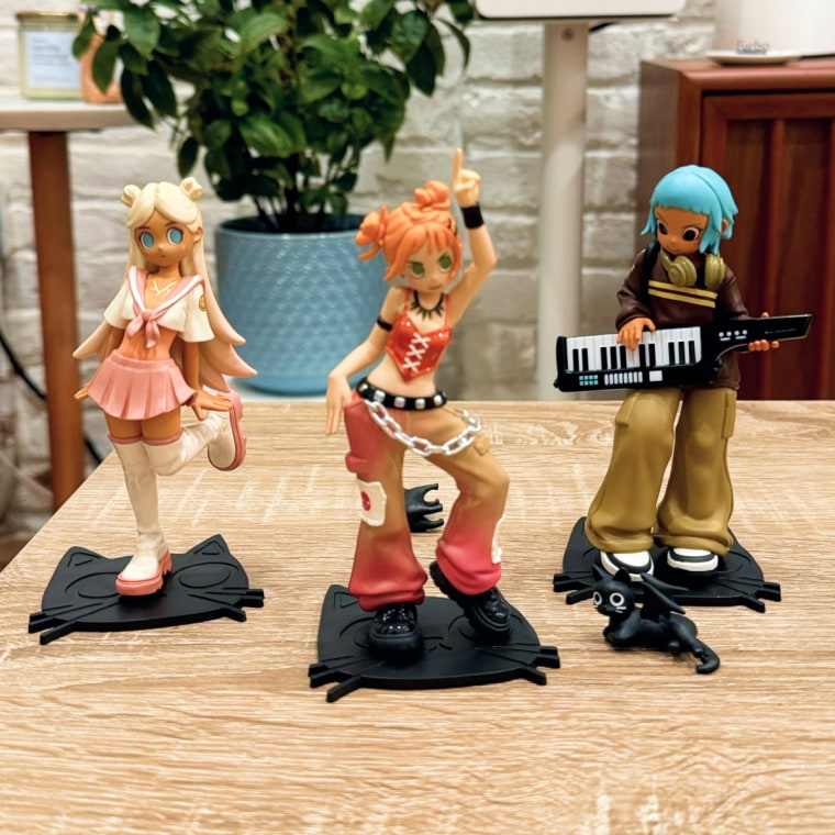 Three Peach Riot Rise Up figurines on a table inf front of a brick wall, a house plant and a speaker.