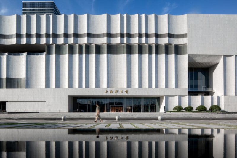 A modern, white building with tall, vertical ridges and a smooth reflective surface in the foreground. A person is walking in front of the building. There's a clear blue sky above.