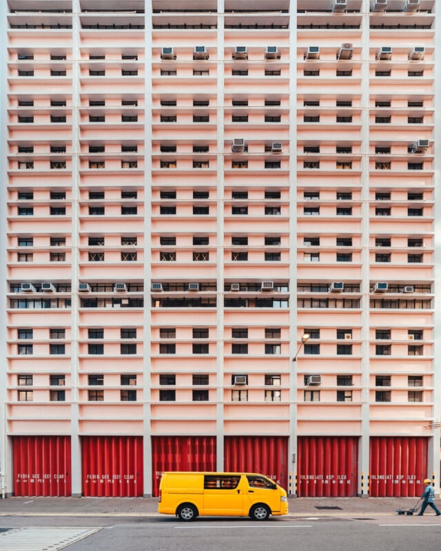 A yellow van is parked in front of a tall building with a repetitive pattern of pink balconies and red garage doors. A person walks past on the sidewalk.