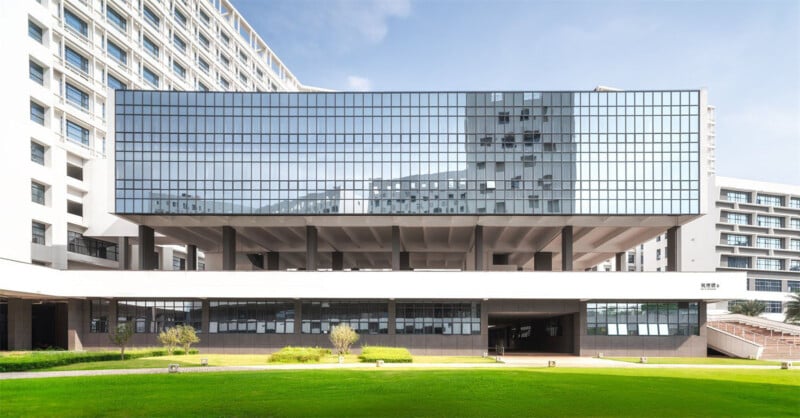 Modern building with a large, overhanging glass facade reflecting surrounding structures. Positioned on a green lawn, the geometric design contrasts with the blue sky. The architecture is sleek and contemporary.