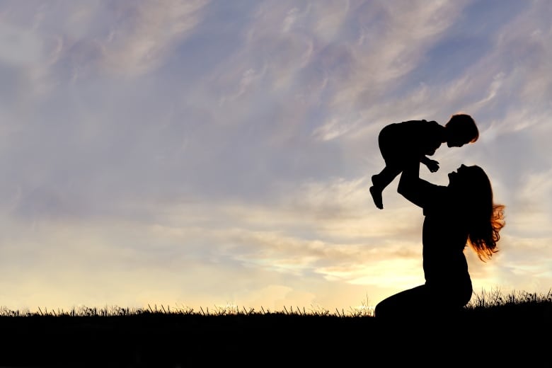 A silhouette of a happy young mother, laughing as she plays with her toddler child and lifts him over her head outside, isolated against the sunset. 